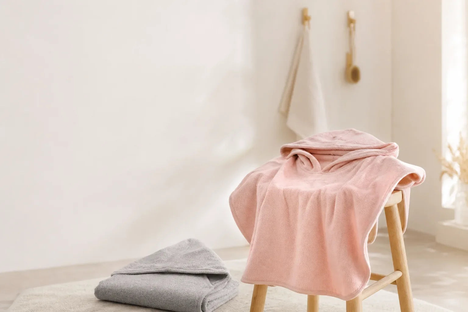 Puderrosa Badeponcho mit Kapuze über einem Holzhocker, daneben ein graues Kapuzenhandtuch