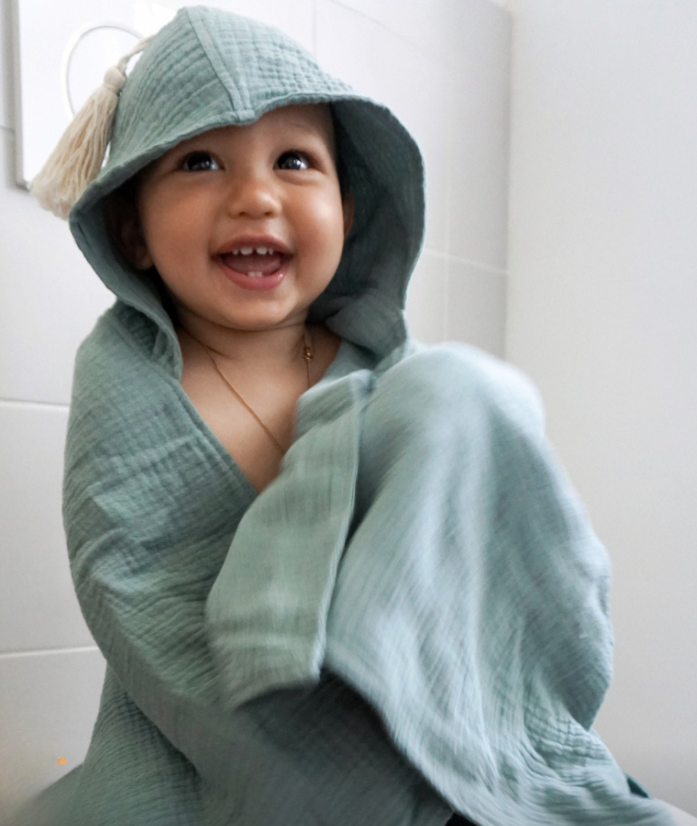 Gemini hat gesagt Ein glücklich lachendes Baby nach einem tränenfreien Bad, eingewickelt in ein weiches, mintgrünes Kapuzenbadetuch mit einer Quaste, als Teil eines entspannten Badeerlebnisses im Abenteuer-Badezimmer.