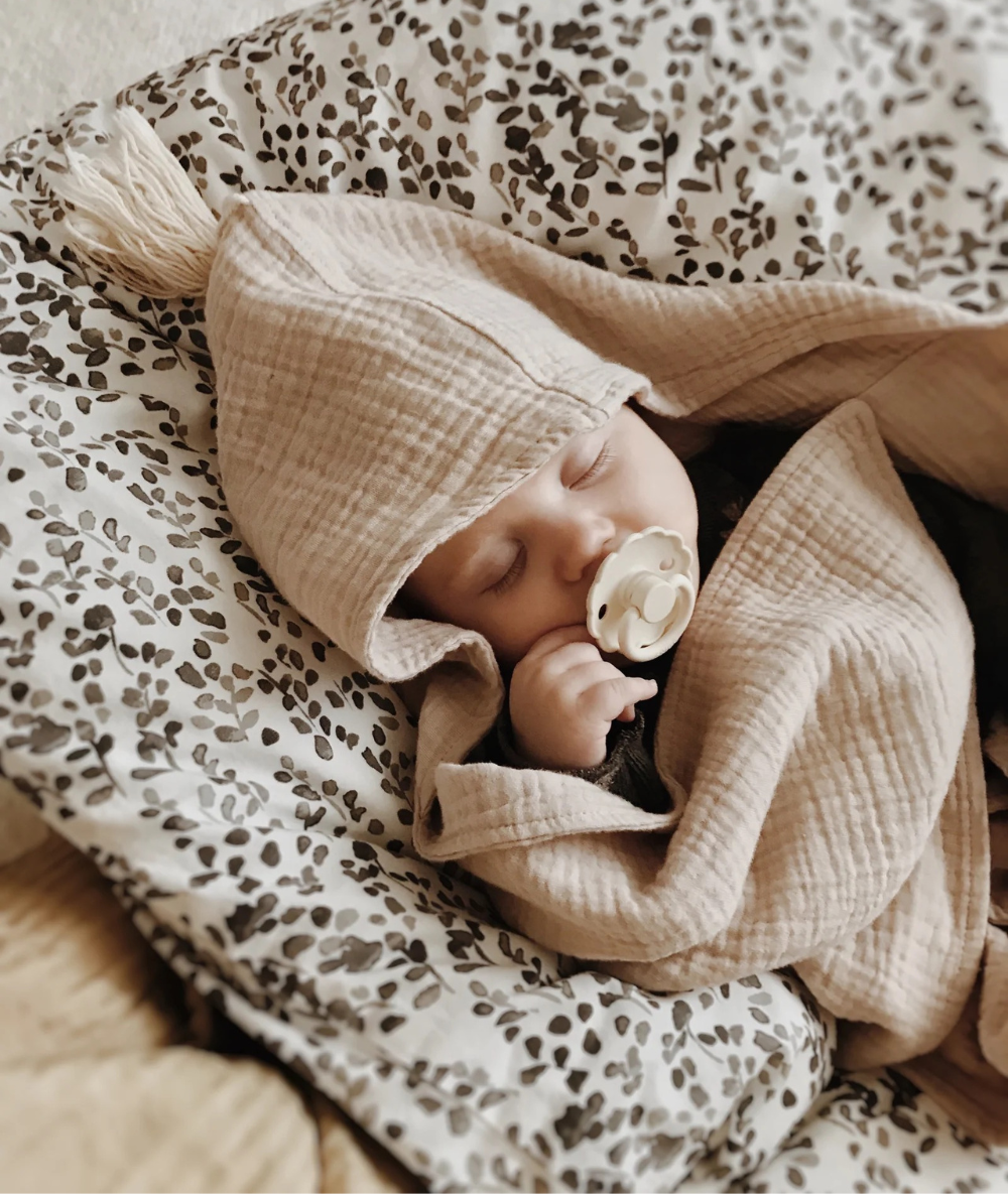 Ein friedlich schlafendes Baby, geborgen eingewickelt in ein Musselin-Kapuzenhandtuch in Naturfarben mit Quaste von Räuberella, als Beispiel für ein einzigartiges, handgefertigtes Baby-Geschenk aus einem kleinen, unabhängigen Shop.