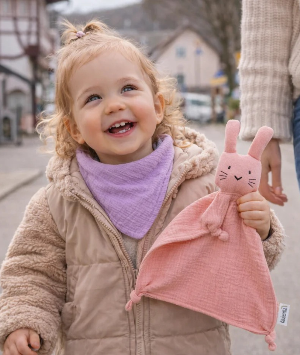 Lachendes Kleinkind beim Frühlingsspaziergang mit fliederfarbenem Musselin-Lätzchen von Räuberella und rosa Hasen-Schmusetuch gegen Sabbern beim Zahnen.