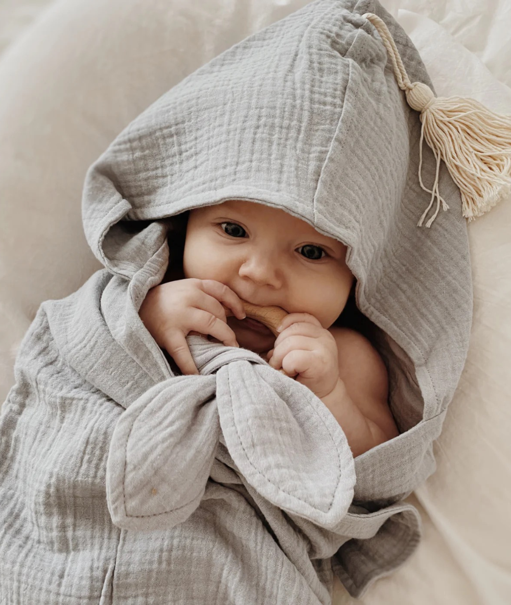 Geborgenes Baby eingekuschelt in ein graues Musselin-Kapuzenhandtuch mit Quaste von Räuberella zum Schutz vor Auskühlung nach dem Baden.